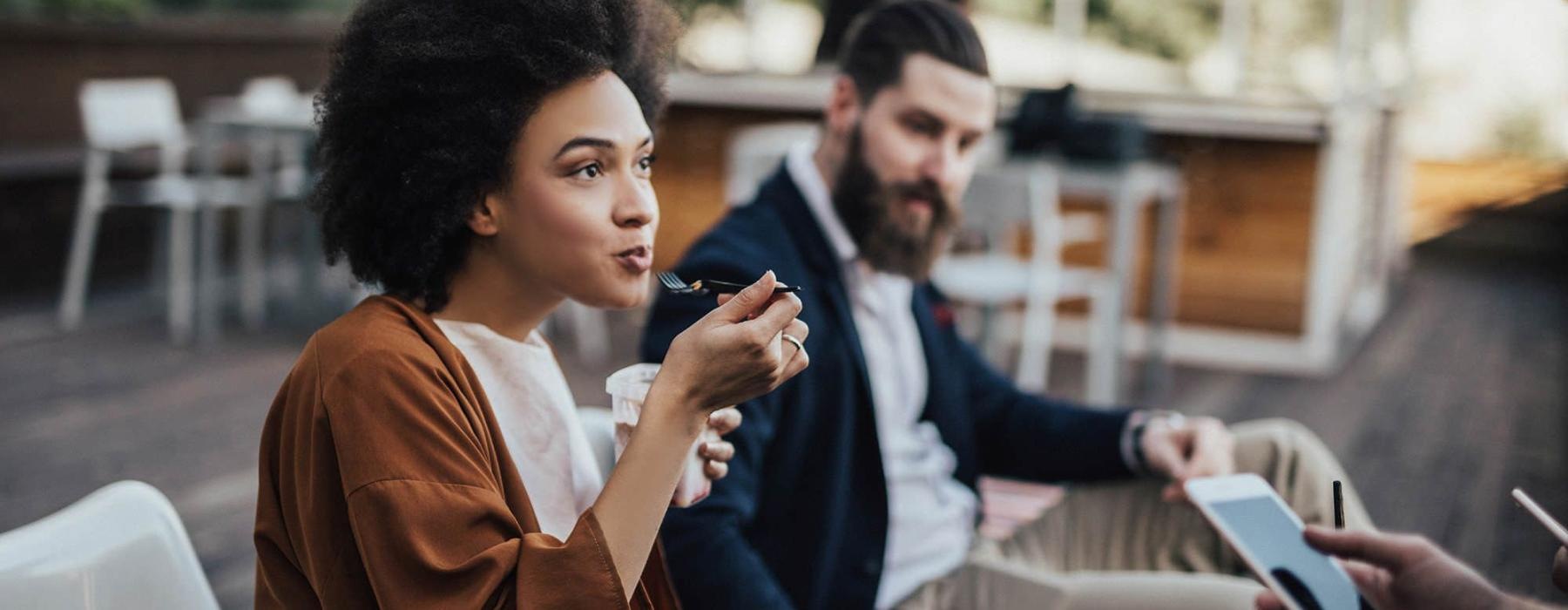 woman sits and eats outside while someone holding a cell talks to her